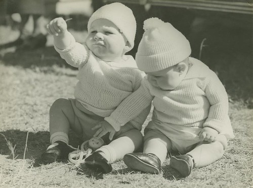 Two little babies sitting on the grass, each wearing caps and warm jumpers