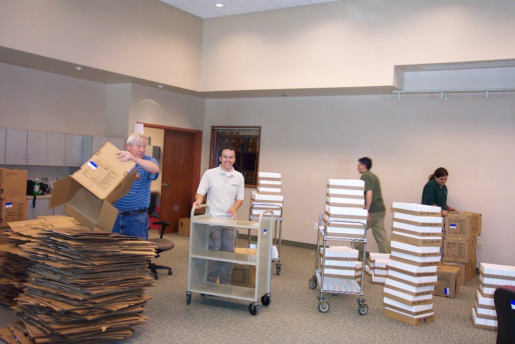 People moving stacks of books and boxes into an empty room.