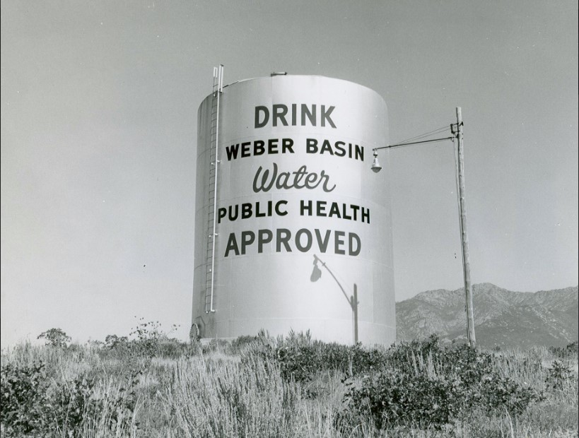 Black and white photo of a large water tower that is painted with the words "DRINK Weber Basin Water Public Health Approved"