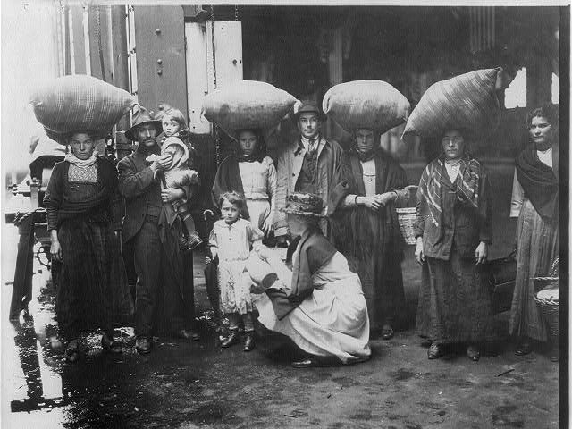 Newly-arrived group of immigrants on pier; women with large sacks balanced on heads; Red Cross woman pouring glass of milk for small girl